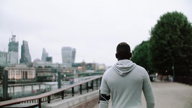 A rear view of young sporty black man runner with smart phone walking on the bridge in a city.