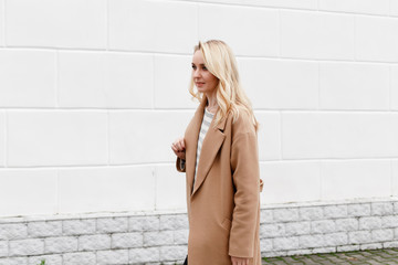 Beautiful young stylish blonde woman wearing long beige coat posing near white street wall. Trendy casual outfit. Street fashion.