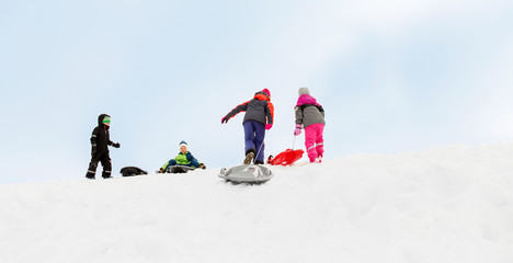 childhood, sledging and season concept - group of happy little kids with sleds climbing snow hill in winter