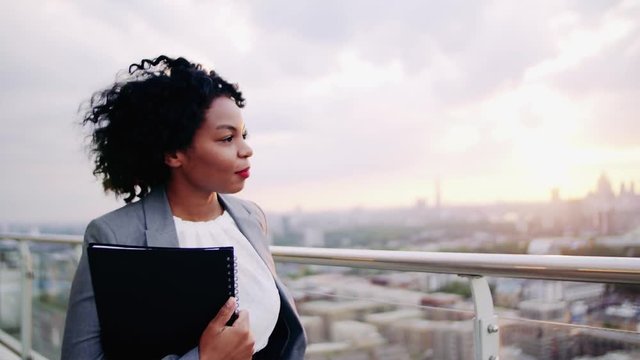 A portrait of a businesswoman walking on a terrace at sunset, holding notes.