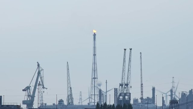 Futuristic scenery with silhouettes of cranes and rotating wind turbines at marine cargo terminal. Gas flare shrouded in mist on background. Sea port of Antwerp, Belgium.