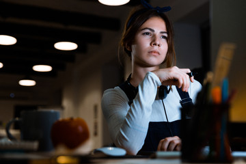 Concentrated young woman designer sitting in office working with tablet and computer at night.