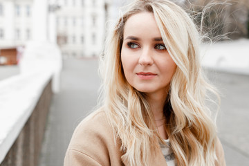 Portrait of beautiful young stylish blonde woman wearing beige coat and walking through the city streets.