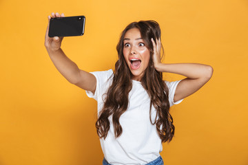 Portrait of caucasian woman 20s with long hair yelling in surprise while taking selfie photo on smartphone, isolated over yellow background