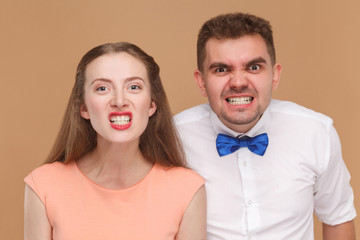 closeup portrait of handsome man and beautiful woman or young couple looking at camera with funny face and showing their clenching teeth. indoor studio shot, isolated on light brown background.
