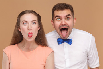 closeup portrait of funny man and beautiful woman or young couple looking at camera with amazed big eyes face and tongue out. indoor studio shot, isolated on light brown background.