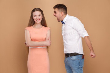 Crazy young man in white shirt standing and screaming at woman in pink dress. woman dont care and looking at camera with toothy smile. indoor studio shot, isolated on light brown background.