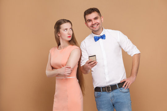 Conflict. Happy man standing, holding mobile phone and looking at camera with toothy smile, and woman looking at him with unhappy face. indoor studio shot, isolated on light brown background.