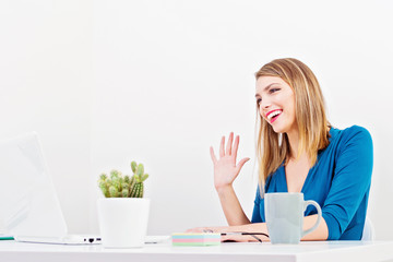 Young woman in her office waving and smiling at her laptop, online communication, chatting or online business meeting concept