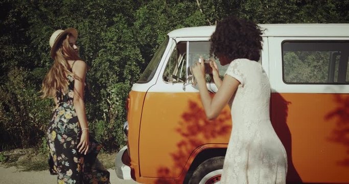 charismatic blonde young woman , wearing a beautiful vintage dresses with a vintage hat , poses for her african curly hair friend who making pictures using a retro camera , beside the orange stylish