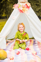 Fototapeta na wymiar Little girl lying and playing in a tent, children's house wigwam in park Autumn portrait of cute curly girl. Harvest or Thanksgiving. autumn decor, party, picnic. Child in halloween costume.