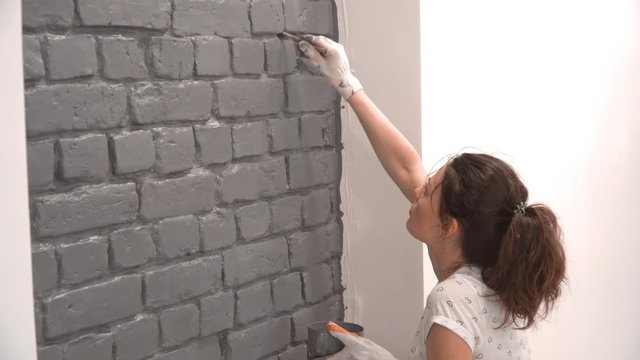 Close-up of a woman painting a brick wall in gray using a paint brush. Renovation in an apartment with white walls.