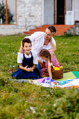 Rear view of father with two children son and daughter paitners in aprons playing together sitting on grass, picnic time over their house on background. The concept of a happy family.