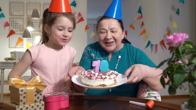 Happy senior woman and her little granddaughter blowing out cake candles to web camera while celebrating birthday with their family via video chat. Slow motion