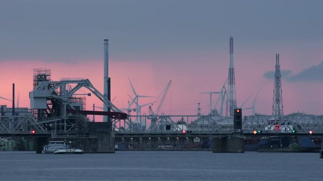 Boat sails under bridge at marine cargo terminal. Futuristic silhouettes view of cranes and rotating wind turbines against beautiful sunset sky on background. Seaport area at evening. Antwerp, Belgium