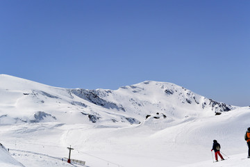 Fototapeta na wymiar White snow-capped mountain slopes with skiers and blue sky with white clouds