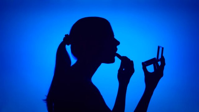 Silhouette of young woman applying lipstick on her lips on blue background. Female's face in profile with mirror. Black contur shadow of teenager's half-face. Concept of fashion and glamour