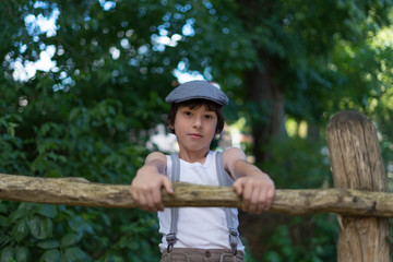 Portrait of a boy dressed in pants with suspenders and a sleeveless shirt near a wooden fence.