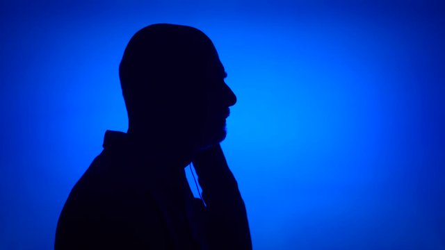 Silhouette of senior man listening to music on smart phone. Male's face in profile putting on ear-phones on blue background. Black contour shadow of grandfather's half-face singing and dancing