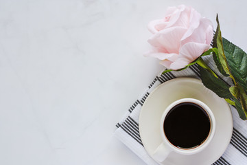 Cup of black coffee with saucer and sweet pink rose on napery against white marble background for drinks and beverage concept