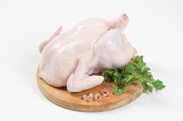 Chicken carcass raw on a round wooden board. On the board next to the chicken is the green of parsley and spices. White background. Close-up. 