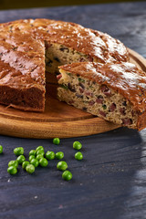 Appetizing unsweetened pie with a salty filling of olives, sausages, meat, vegetables, peas. A handful of green peas as a decoration in the foreground. Big pie lies on a beautiful wooden round board.