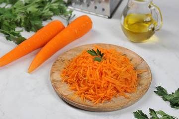 Juicy grated carrots on a wooden tray. Next to a few carrots, parsley and a jug of olive oil. Light background. Close-up.