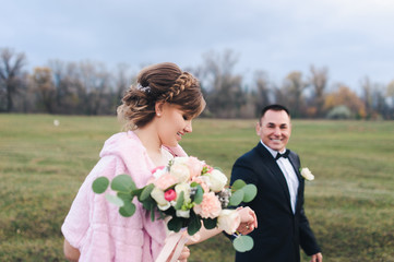 An adult groom and pretty bride strolling along the green field against the backdrop of a pine forest and smiling. Wedding portrait close-up of beautiful newlyweds.