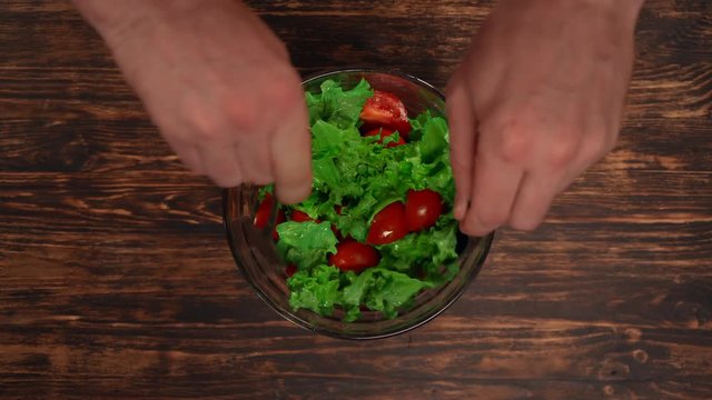 man is mixing green-stuff in a bowl. lettuce leaves with tomatos in a plate on wooden table.