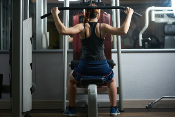 Fototapeta na wymiar Young fit woman flexing muscles on gym machine in front of a mirror. Sport, fitness, strength training