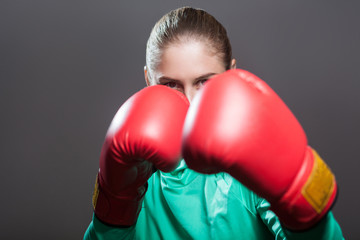 Portrait of confident beautiful young athlete woman with collected hair standing on the defensive in boxing red gloves. Indoor studio shot, isolated on dark gray background.