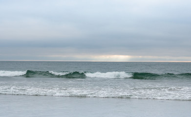 Fototapeta na wymiar Wave rolling in towards shore, soft light blue clouds, sunshine on horizon