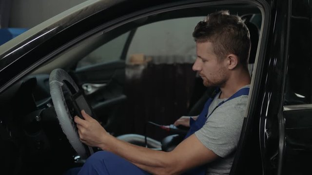 handsome mechanic in uniform is working in auto service. Car repair and maintenance. Performing engine diagnostics with tablet pc laptop