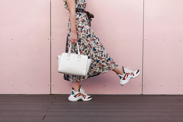 Stylish beautiful young woman in fashionable dress with stylish white bag and trendy sneakers is...