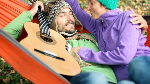 Happy couple chilling in hammock in autumn forest. They kiss, smile and talk. Guitar lying nearby.