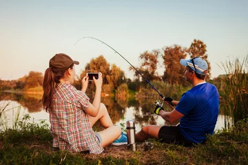 Poster Jong koppel vissen en thee drinken op de rivier bij zonsondergang. Vrouw filmt haar vriend die vis vangt. Mensen die plezier hebben © maryviolet