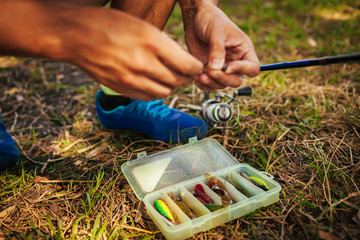 Young man attaching bait to hook of spinning. Fishing tackle. Man chooses lure in fishing gear box.