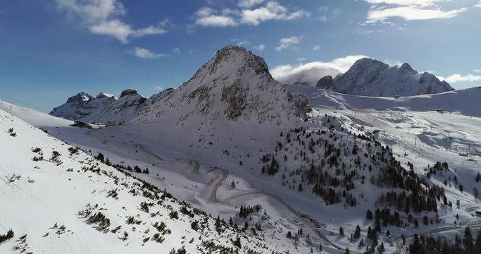 Forward aerial over snowy valley with woods forest and hairpin bend road at Pordoi pass. Sunny day with cloudy sky.Winter Dolomites Italian Alps mountains outdoor nature establisher.4k drone flight