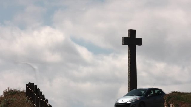 Zooming in timelapse of a village stone cross memorial with beautiful blue sky and clouds moving in the background. In the foreground, various vehicles move along the road. Budleigh Salterton