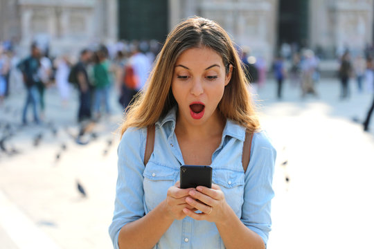 Surprised young woman using smart phone outdoors. Close up portrait surprised screaming girl looking at phone seeing news or photos with funny emotion on her face isolated outside city background.