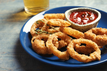 Plate with homemade crunchy fried onion rings and tomato sauce on wooden background, closeup