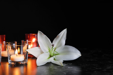 White lily and burning candles on table in darkness, space for text. Funeral symbol