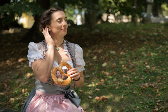 Woman wearing a traditional German dirndl sitting in park eating a pretzel