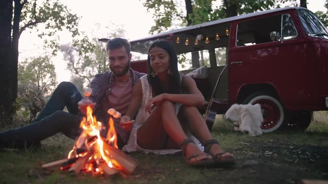 Beautiful young couple roasting marshmallows over a campfire while enjoying their road travel, slow motion, dolly