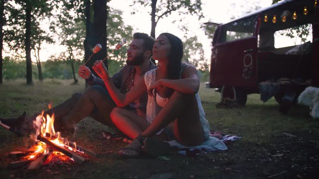 Beautiful young couple roasting marshmallows over a campfire while enjoying their road travel, slow motion, dolly