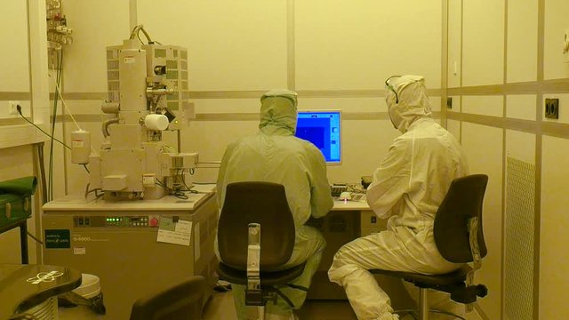A shot of the back of two scientists working together on a big electrone microscope in the clean room.