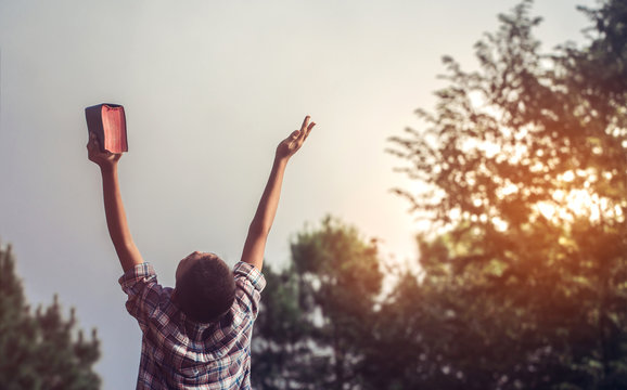 Boy holding the Holy Bible and lift hand for worship God with ligtf of sunset in mountain background.