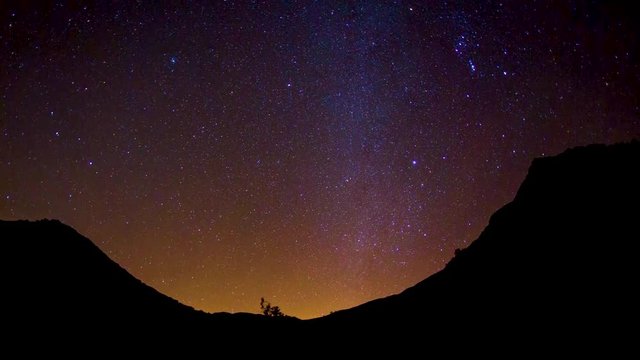 timelapse of stars in the orange sky moving while a silhouette of a mountain sits in the foreground