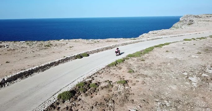 Italian couple riding scooter on the road in the island of Lampedusa, Italy during summer vacation with Mediterranean sea in background