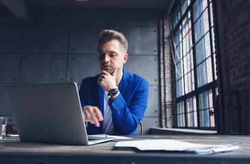 Young attractive business man in blue suit is sitting in the office at the desk with laptop and...